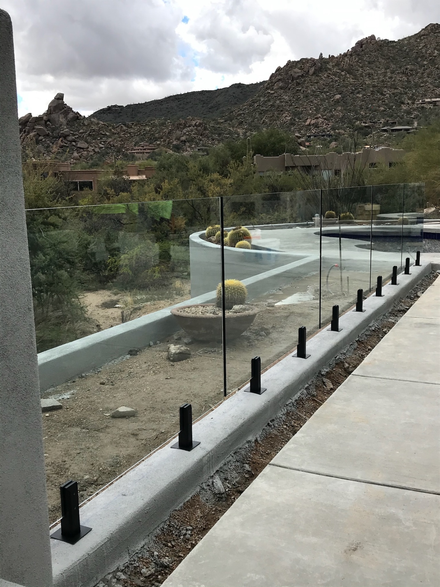 Long run of glass fence panels with black spigots set on a painted bond beam, framing the desert pool deck and granite mountain backdrop in Arizona.
