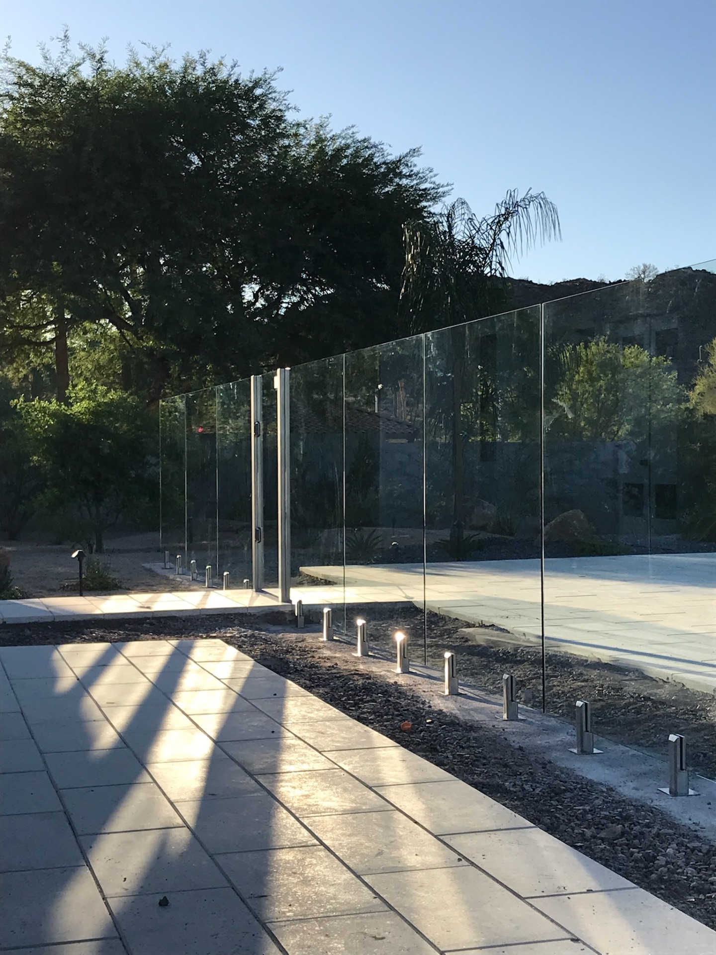 Long line of frameless glass fencing and brushed stainless self-closing gate hardware illuminated by paver deck up-lighting on an Arizona estate.