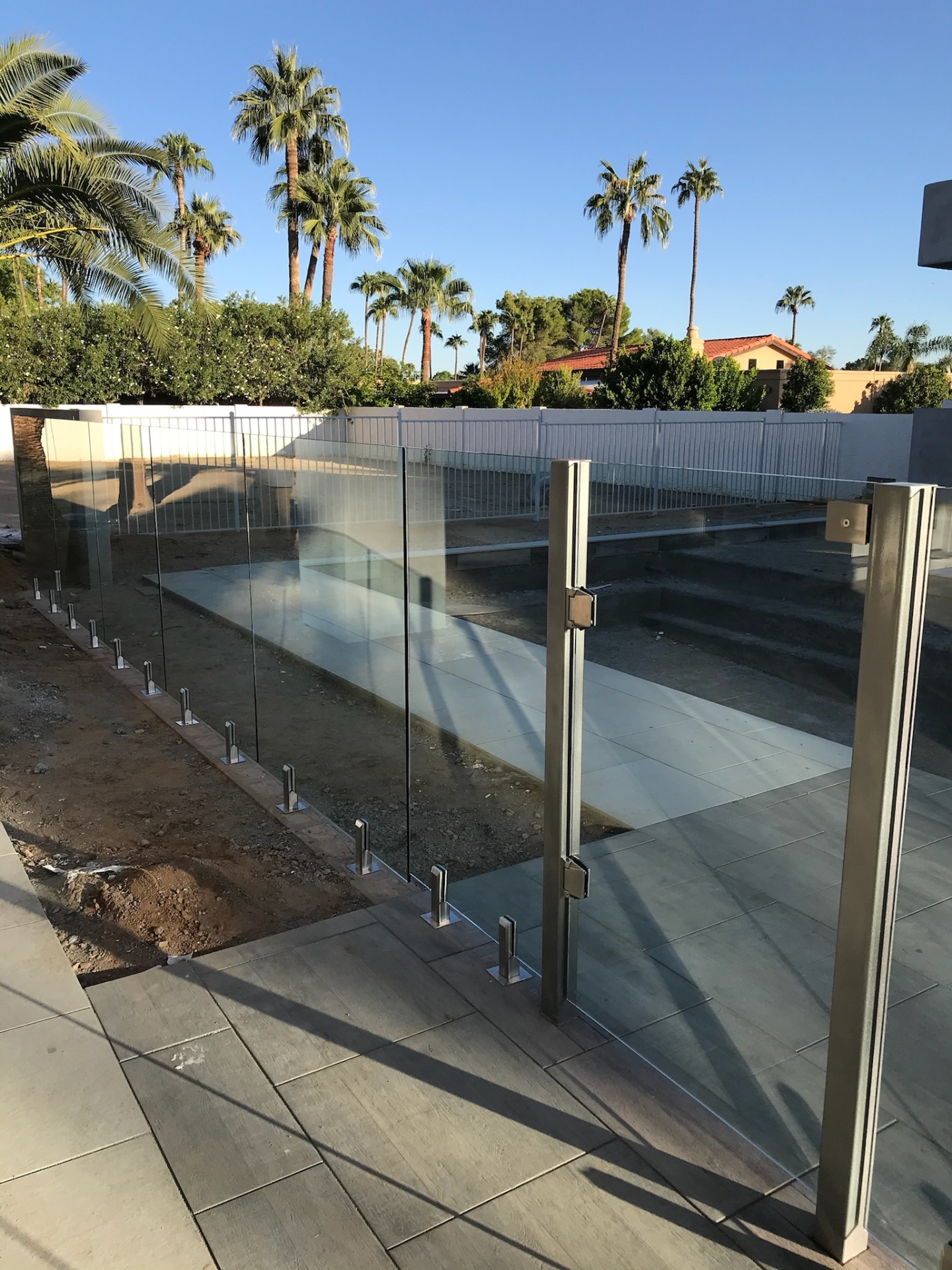 Frameless glass fencing and stainless gate framing a new pool deck — code-height tempered glass fence panels on spigot mounts with palms and white perimeter fence beyond in Phoenix.