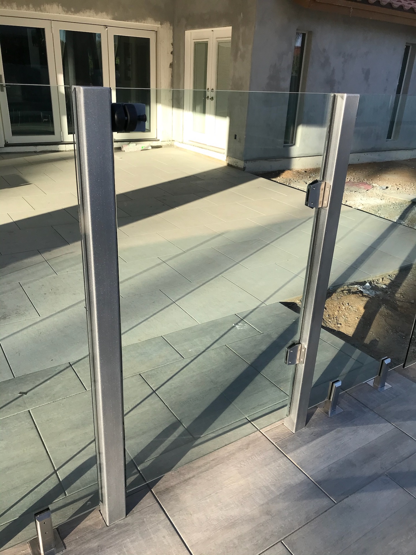 Stainless gate posts and hinged frameless glass fence gate panel set on porcelain pavers — close detail of Sonoran Glass fencing spigot, hinge, and latch hardware.