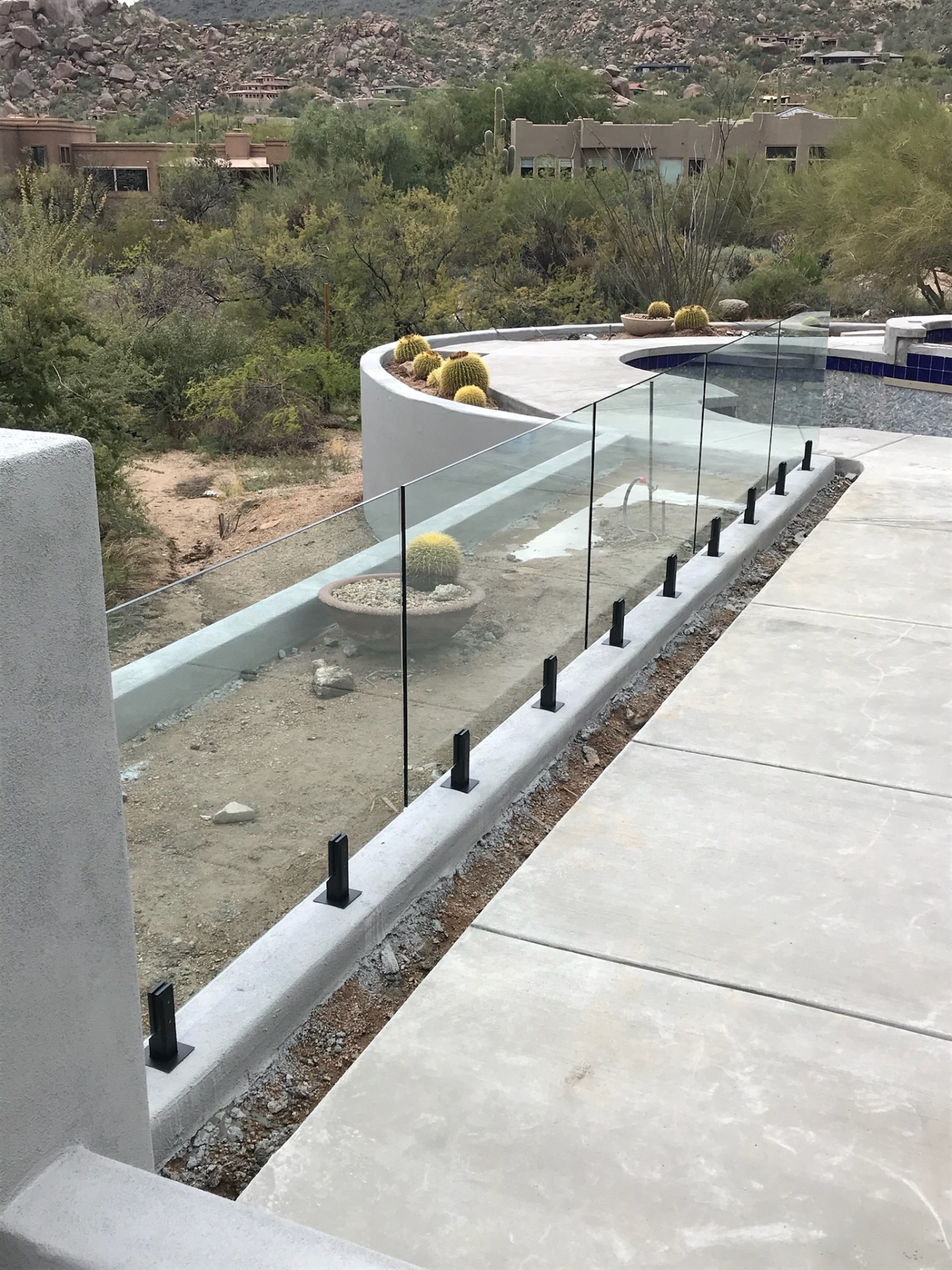 Curved glass fence on a freshly poured concrete bond beam, boulder hillside and barrel cactus planters beyond — North Scottsdale glass fencing install.