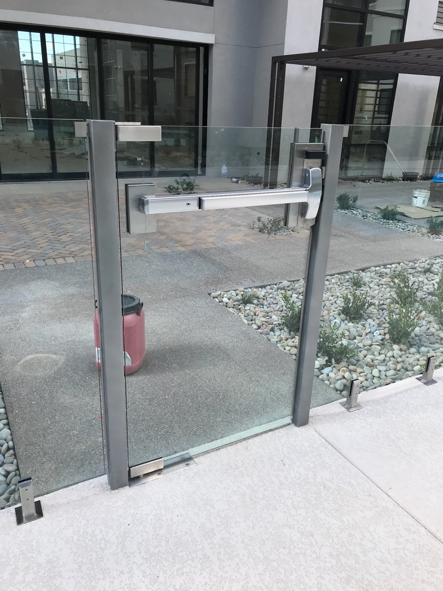 Close view of a multifamily glass fence gate with aluminum framing, panic bar hardware, and adjacent tempered glass fence panels.